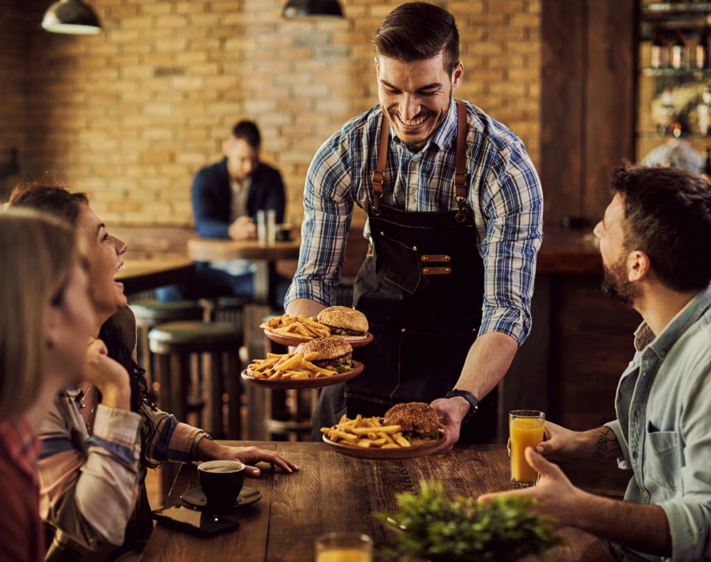 Group of happy friends having fun while waiter is serving them food in a pub. Focus is on waiter.